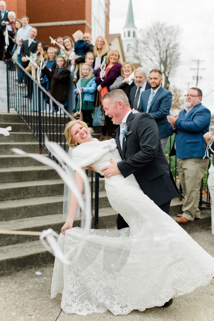 A bride and groom laughing on church steps as guests wave ribbon wands