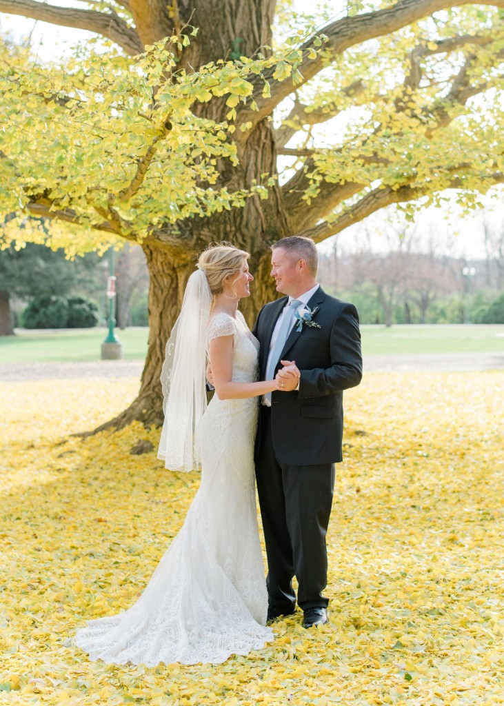 A bride and groom holding hands beneath a ginkgo tree in a carpet of yellow leaves