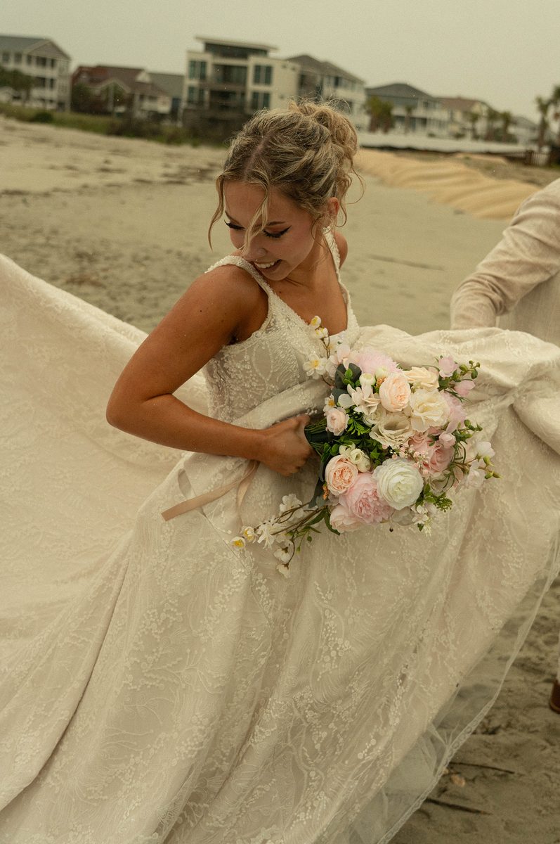 A bride in a lace gown laughing on a beach, holding a bouquet of pink and white roses