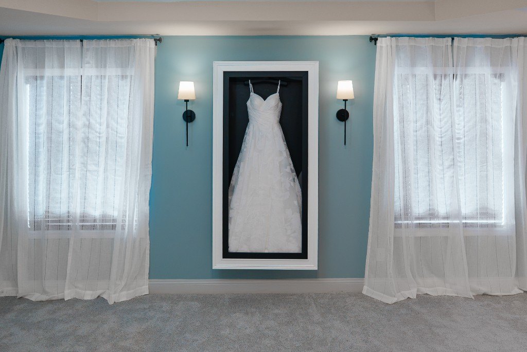 A wall-mounted Mrs. Case Gallery displaying a lace gown on a blue wall between two windows