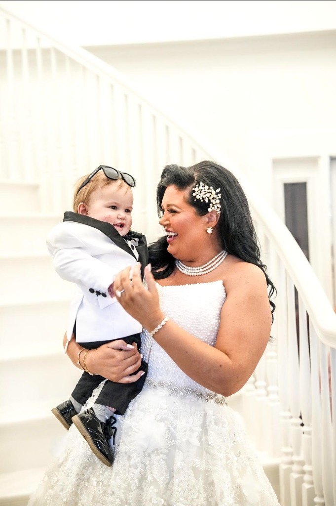 A bride in a beaded gown holding a child in a white jacket on a staircase