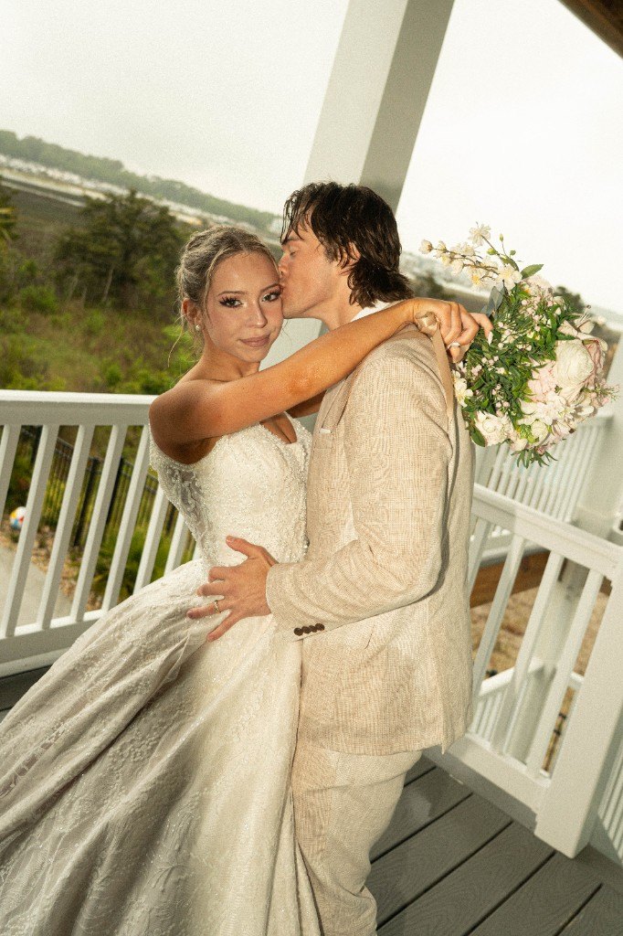 A groom kissing his bride on a porch, her lace gown and bouquet caught in the afternoon light